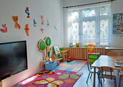 Pokój dziecięcy z kolorowym dywanem, stolikami, krzesłami, zabawkami i dekoracjami ściennymi.