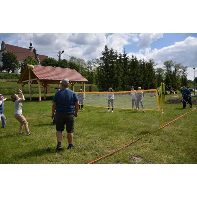 Grupa osób grających w siatkówkę na trawiastym boisku, w tle altana, budynek i drzewa.