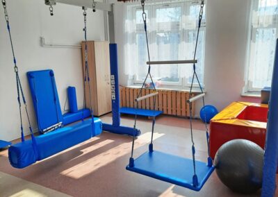Pokój terapeutyczny z huśtawkami sensorycznymi, matami, piłkami i dużymi oknami.