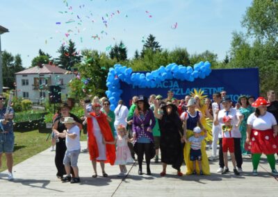 Ludzie w baśniowych kostiumach na zewnątrz, z niebieskimi balonami i konfetti w tle.