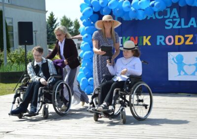 Piknik rodzinny, dwie osoby na wózkach inwalidzkich z opiekunami, niebieskie balony w tle.