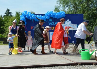 Grupa osób w kostiumach na drewnianym podeście z niebieskimi balonami i napisem "Fundacja Szlachta Pisaków".