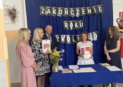 Grupa ludzi z dyplomami na zakończeniu roku szkolnego przed niebieskim tłem z kwiatami na stole