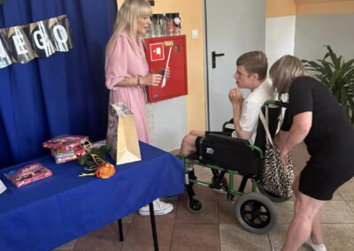 Kobieta w różowej sukience rozmawia z chłopcem na wózku; stół z prezentami, dekoracje, sprzęt gaśniczy.