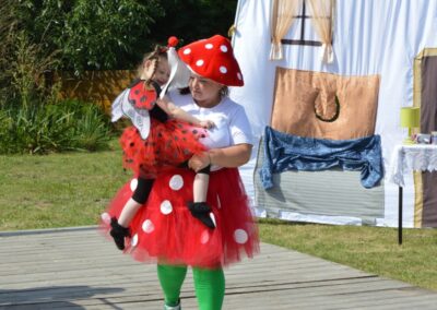 Dwie osoby w kostiumach grzyba i biedronki na drewnianej platformie na tle dekoracji teatralnej.