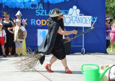 Osoba przebrana za wiedźmę na scenie, w tle inne osoby w kostiumach i baner "Fundacja Szlaki".