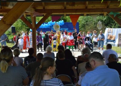 Grupa ludzi w kolorowych kostiumach na scenie podczas wydarzenia plenerowego z balonami w tle.