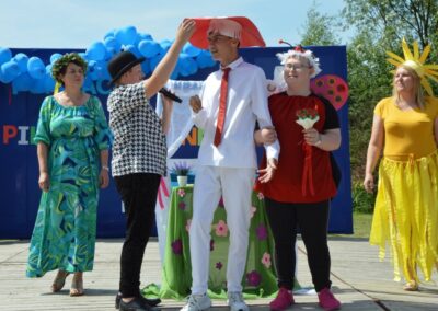 Osoby w kostiumach na scenie z balonami, stołem z kwiatami i kolorowym tłem.