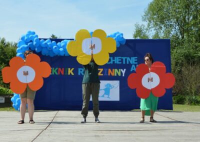 Trzy osoby trzymające duże, kolorowe kwiaty na scenie z ozdobami i napisem "Piknik Rodzinny".