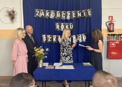 Sala szkolna podczas zakończenia roku szkolnego, osoby przy stole z niebieskim obrusem, napisy w tle