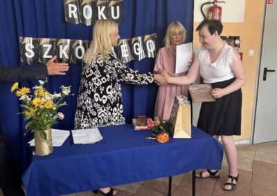 Ceremonia zakończenia roku szkolnego, wręczanie dyplomu uczennicy przy stole z dekoracjami.