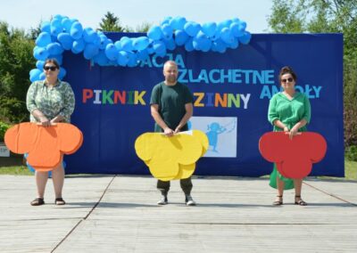 Troje ludzi na scenie z kolorowymi tekturowymi figurami i niebieskim banerem "Fundacja Szlachetne Anioły Piknik Rodzinny".