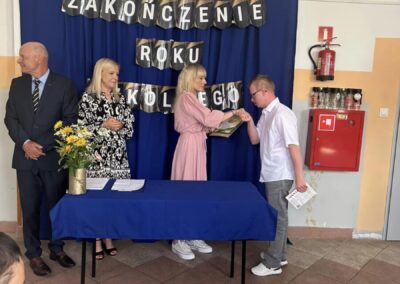 Uroczystość zakończenia roku szkolnego, uczeń odbiera nagrodę od kobiety przy stole z niebieskim obrusem