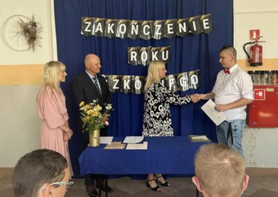 Uroczystość zakończenia roku szkolnego, osoby przy stole z dokumentami.