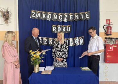 Cztery osoby stojące przy stole z kwiatami i dokumentami, napis "Zakończenie roku szkolnego".