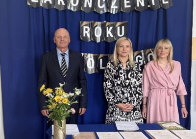 Grupa trzech osób na tle zasłony z napisem "Zakończenie Roku Szkolnego" przed stołem z dyplomami.