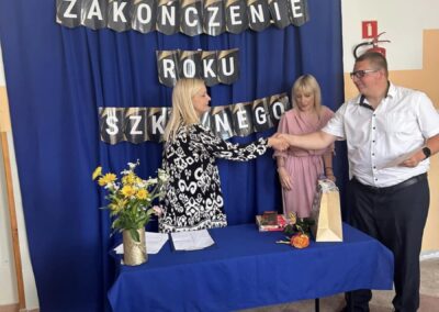 Zakończenie roku szkolnego, kobieta podaje rękę mężczyźnie przy stole z kwiatami i dokumentami.