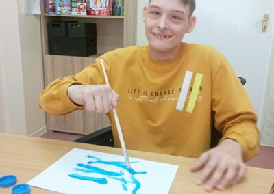 Chłopiec w żółtej bluzie siedzi przy stole, malując na kartce papieru niebieską farbą. Uśmiecha się, trzymając pędzel w ręku.