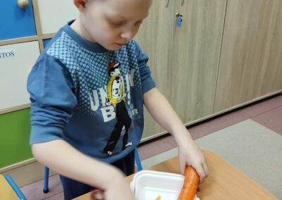 Chłopiec w niebieskiej bluzie z krótkimi włosami obiera dużą marchewkę nad plastikowym pojemnikiem. Tło stanowią szafki z drewnianymi drzwiami.