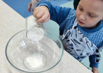 Chłopiec w niebieskiej bluzie przesypuje mąkę z kubka do dużej przezroczystej miski. W tle znajduje się dziewczynka w różowej koszulce z okularami, zajmująca się swoimi zadaniami.