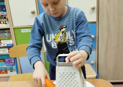 Chłopiec w niebieskiej bluzie trze marchewkę na metalowej tarce nad papierowym talerzem. W tle widoczne są kolorowe półki i szafki.