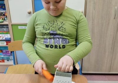 Chłopiec w zielonej koszulce z nadrukiem "Dino World" trze marchewkę na metalowej tarce nad papierowym talerzem. Za nim widoczne są półki z książkami i szafki.