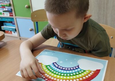 Chłopiec siedzi przy stole i pracuje nad kartką papieru z tęczą, na którą przykleja kolorowe naklejki w kształcie kropek. W tle widoczne są kolorowe półki z różnymi materiałami edukacyjnymi.