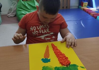 Chłopiec w czerwonej koszulce siedzi przy stoliku, wyklejając marchewkę z kolorowego papieru na żółtej kartce. W tle widoczne są dekoracje w oknie oraz kolorowe poduszki.