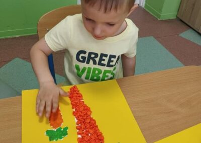 Chłopiec w jasnej koszulce z napisem "Green Vibes" siedzi przy stoliku, wyklejając kolorowy obrazek na żółtej kartce. Skupia się na swojej pracy.