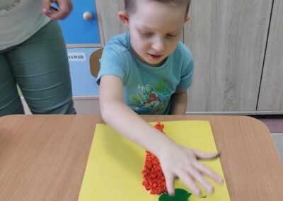 Chłopiec w niebieskiej koszulce siedzi przy stoliku, wyklejając zieloną farbą na żółtej kartce. Obok niego stoi osoba dorosła pomagająca mu w pracy.