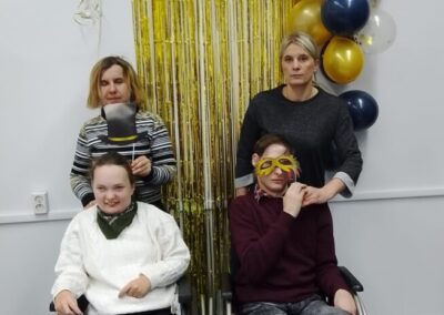 Dwie osoby na wózkach inwalidzkich pozują na tle złotej kurtyny z balonami. Za nimi stoją dwie kobiety, trzymające rekwizyty maski i kapelusza.