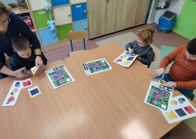 Trójka dzieci siedzi przy stole, wycinając kolorowe obrazki z papieru. Jedno z dzieci otrzymuje pomoc od osoby dorosłej, która stoi obok. W tle widoczne są półki z zabawkami i kolorowymi szafkami.