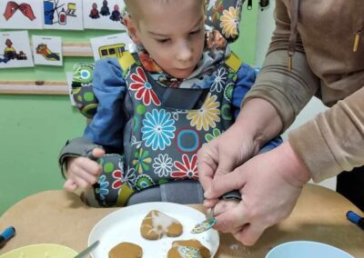 Mały chłopiec, siedzący na specjalnym foteliku z kolorowym wzorem kwiatów, obserwuje, jak nauczyciel pomaga mu dekorować pierniki na talerzu. Chłopiec jest skupiony, a na stole widać różne akcesoria do dekorowania, w tym miseczki z posypką i lukrem.