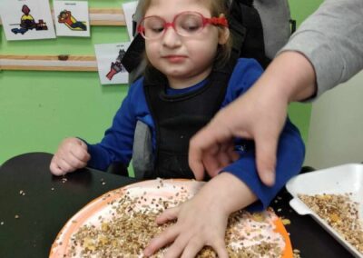 Dziewczynka w okularach, ubrana w niebieską bluzę, pracuje nad projektem, na którym przykleja ziarna na okrągłej kartce papieru. Nauczyciel pomaga jej w pracy, delikatnie prowadząc jej rękę. W tle widać plansze edukacyjne z obrazkami, co sugeruje, że praca odbywa się w szkole lub przedszkolu.