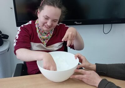 Dziewczynka w bordowej bluzie siedzi przy stole, wkładając rękę do miski z białą masą. Jest uśmiechnięta, a obok niej widoczne są ręce innej osoby, która trzyma miskę.