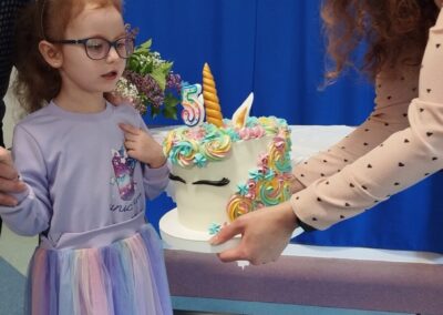 Dziewczynka w fioletowej sukience i okularach stoi przy stole z tortem udekorowanym motywem jednorożca. Obok niej dorosła osoba pomaga ustawić tort. W tle widoczne są dekoracje urodzinowe z napisem "Tosia".