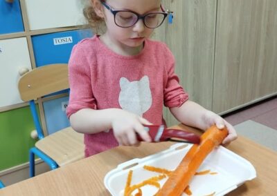 Dziewczynka w różowej bluzce i okularach obiera marchewkę nad plastikowym pojemnikiem. Siedzi przy stoliku, a w tle znajdują się szafki z kolorowymi drzwiami.