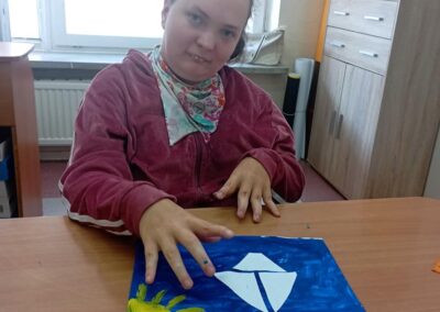 Dziewczynka w bordowej bluzie siedzi przy stole, pracując nad plastyczną pracą przedstawiającą białą łódkę na niebieskim tle z żółtym słońcem.