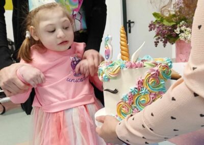 Mała dziewczynka w różowej sukience i urodzinowej czapeczce stoi przy stole, na którym znajduje się pięknie ozdobiony tort w kształcie jednorożca. Dziewczynka patrzy na tort, a osoby dorosłe pomagają jej w świętowaniu urodzin.