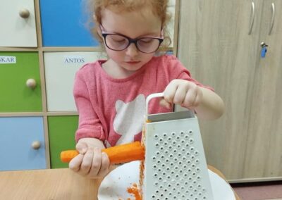 Dziewczynka w różowej bluzce i okularach trze marchewkę na metalowej tarce nad papierowym talerzem. Siedzi przy stole, a w tle widoczne są szafki z kolorowymi drzwiami.