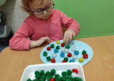 Dziewczynka w różowej bluzie siedzi przy stole i układa na kartce kolorowe pompony. W tle widać plakaty z obrazkami, które mogą przedstawiać różne czynności lub przedmioty.