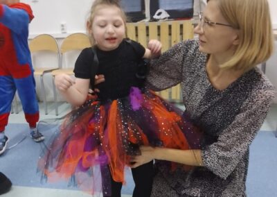 Mała dziewczynka ubrana w czarno-pomarańczowy kostium wiedźmy stoi obok nauczyciela, który pomaga jej poprawić strój. Dziewczynka ma na głowie czarny, spiczasty kapelusz. W tle widać inne dzieci w przebraniach, co sugeruje, że odbywa się zabawa tematyczna.