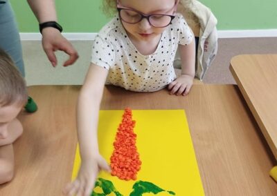 Dziewczynka w białej bluzce w kropki i okularach wykleja marchewkę na żółtej kartce. Obok niej dorosła osoba pomaga jej w pracy. W tle widoczna jest zielona ściana.