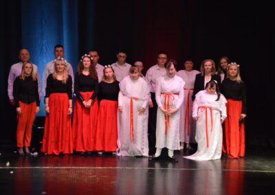 Grupa osób, w tym kobiety i mężczyźni, pozuje na scenie podczas występu. Część osób ubrana jest w tradycyjne stroje ludowe - czerwone spódnice i czarne bluzki, a inne osoby mają na sobie białe stroje z czerwonymi wstążkami. Kilka kobiet ma na głowach kwieciste wianki. Tło sceny jest ciemne, co podkreśla oświetlenie skierowane na uczestników. Wyraz twarzy uczestników sugeruje, że są zaangażowani w występ artystyczny lub teatralny.