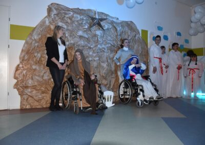 Grupa osób przebrana w stroje jasełkowe, w tym dwie osoby na wózkach inwalidzkich, uczestniczy w przedstawieniu bożonarodzeniowym. W tle widoczna jest dekoracja imitująca grotę.