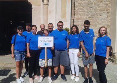 Grupa osób, ubrana w niebieskie koszulki z logo „Szlachetne Anioły”, pozuje do zdjęcia przed wejściem do kamiennego kościoła. Na środku jedna z osób trzyma tabliczkę z napisem „Fundacja Szlachetne Anioły w Klimontowie”. Tło stanowi kamienna fasada kościoła, a pogoda jest słoneczna, co sugeruje ciepły, letni dzień. Uczestnicy wyglądają na zadowolonych i dumni z reprezentowania swojej organizacji.