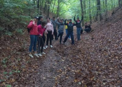 Grupa uczniów i nauczycieli podczas spaceru w lesie, otoczona drzewami, na ścieżce pokrytej liśćmi.
