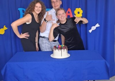 Trzy osoby z grupy, ubrane w czarne ubrania, stoją przed stołem z tortem urodzinowym, na tle dekoracji z napisem "Urodziny". Wszyscy uśmiechają się do kamery.