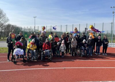 Grupa osób, w tym dzieci, dorośli oraz osoby na wózkach inwalidzkich, pozuje do zdjęcia na bieżni sportowej podczas plenerowej imprezy. Część osób trzyma kolorowe ozdoby w kształcie kwiatów, biedronek i innych wiosennych motywów. W tle widoczne są strachy na wróble ozdobione różnokolorowymi materiałami. Dookoła rozpościera się przestrzeń z drzewami i ogrodzeniem sportowym, a niebo jest bezchmurne, co sugeruje słoneczną pogodę.