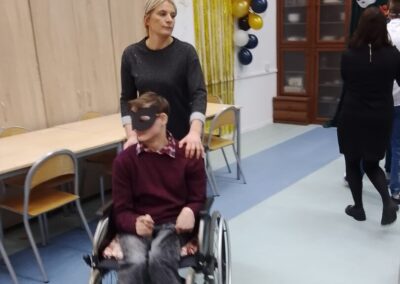 Kobieta prowadzi chłopca na wózku inwalidzkim po sali ozdobionej girlandami i balonami. Chłopiec ma na sobie maskę karnawałową.